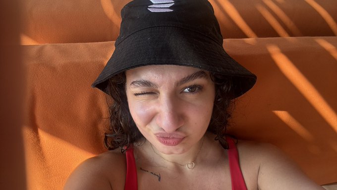 A young woman winks and makes a playful face while wearing a black bucket hat with a logo, photographed against a warm orange backdrop.