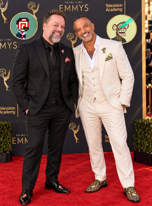 Two men posing together on the red carpet at the Prime Time Emmys Television Academy event; the man on the left wears a black suit, while the man on the right wears a white suit with snake-print shoes.
