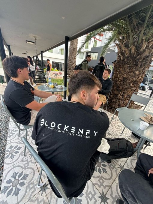 Outdoor gathering at a patio with palm trees, where attendees sit at tables; one person wears a black t-shirt with 'BLOCKENFY - Tu mejor opción para tokenizar' printed on the back.