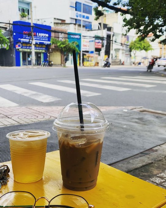 A street-level photograph showing two iced beverages in plastic cups—one light-colored and one dark iced coffee with a straw—sitting on a yellow table at a sidewalk café overlooking a city street with shops and motorbikes.