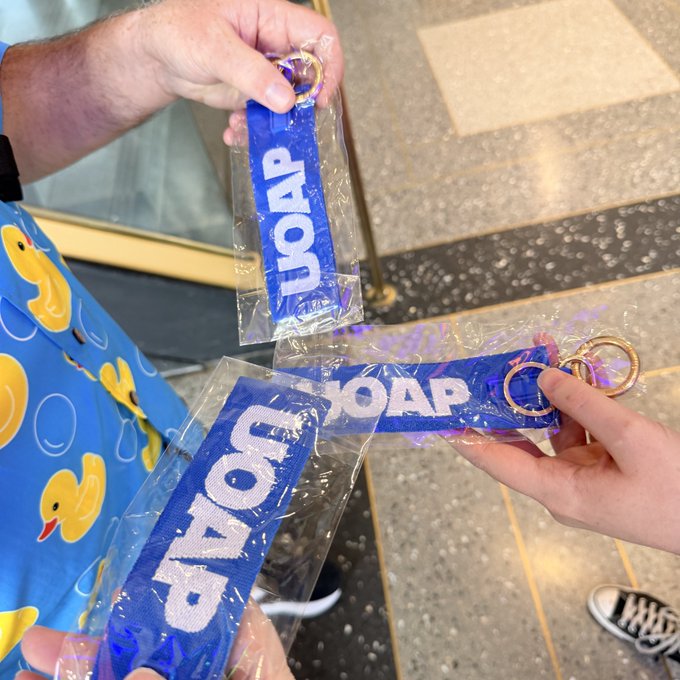 Hands holding up clear plastic packages containing purple lanyards and keychains with the word "SOAR" printed in white letters.