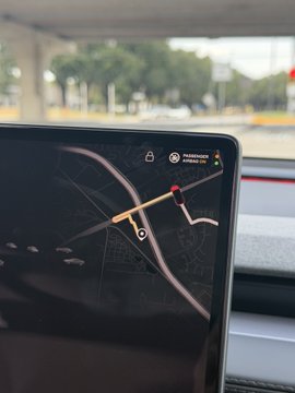 A vehicle's dashboard navigation screen displaying a map with a route highlighted, with a road and trees visible through the windshield.