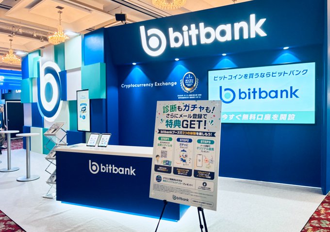 The image shows the bitbank cryptocurrency exchange booth, featuring a blue and white design with the prominent "bitbank" logo, a "Cryptocurrency Exchange" label, and a Japanese promotional poster about a referral campaign offering diagnostic gadgets ("診断もガチャも特典GET!"). The booth display, which includes iPads and informational signage, reflects bitbank's presence at WebX2026, where they engaged with attendees despite the hot weather.