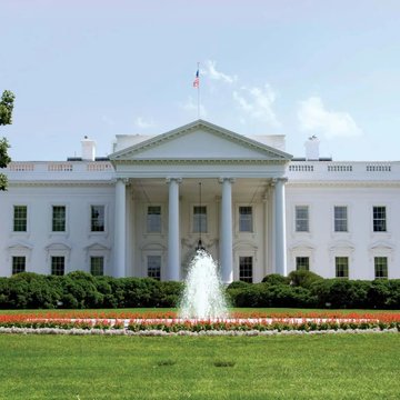 The White House is shown on a sunny day with a flag flying above its columned portico and a fountain spraying in the manicured front garden. The image relates to the post by providing a visual reference to the White House, the institution referenced in the headline about advancing the CLARITY Act.