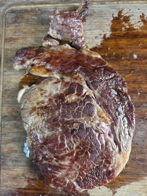A cooked, well-seared steak rests on a wooden cutting board, its juices pooling around it.