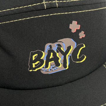 A black cap with yellow contrast stitching features a "BAYC" (Bored Ape Yacht Club) graphic with an ape skull design, matching the official tee from the BAYC x Stonrok Anchor Drop pop-up collection available in Seoul.