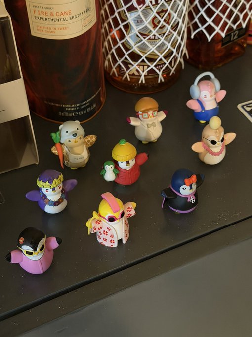 A collection of colorful Pudy Penguins figurines arranged on a dark surface next to bottles of "Fire & Cane" experimental series rum from Westland Distillery. The adorable chibi-style characters feature various themed outfits, from a chef and pumpkin-headed figure to one holding a carrot, perfectly illustrating the post's excitement about wanting to collect the entire set.