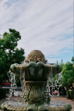 A close-up photograph of an ornate green and gold fountain with water spraying upward, set against a backdrop of trees and a cloudy blue sky.