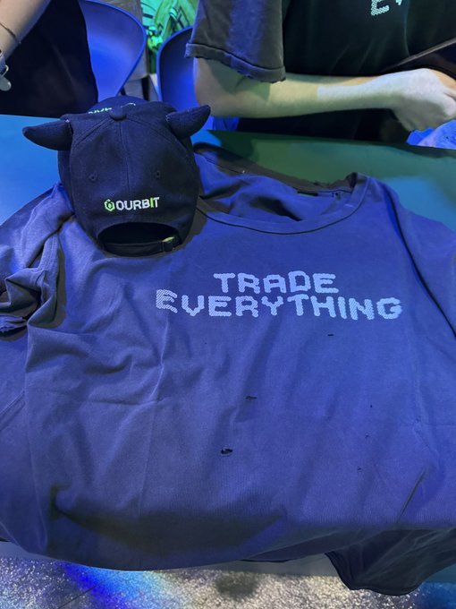 A blue t-shirt with 'TRADE EVERYTHING' in pixelated text sits next to a navy cap featuring a 'OURBIT' logo.