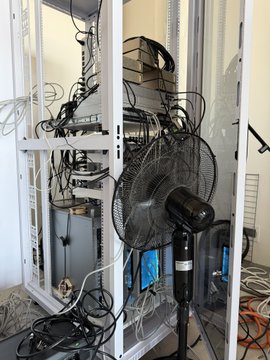 An equipment rack with tangled cables, electronic devices, and a black standing fan pointed at it, likely for cooling.