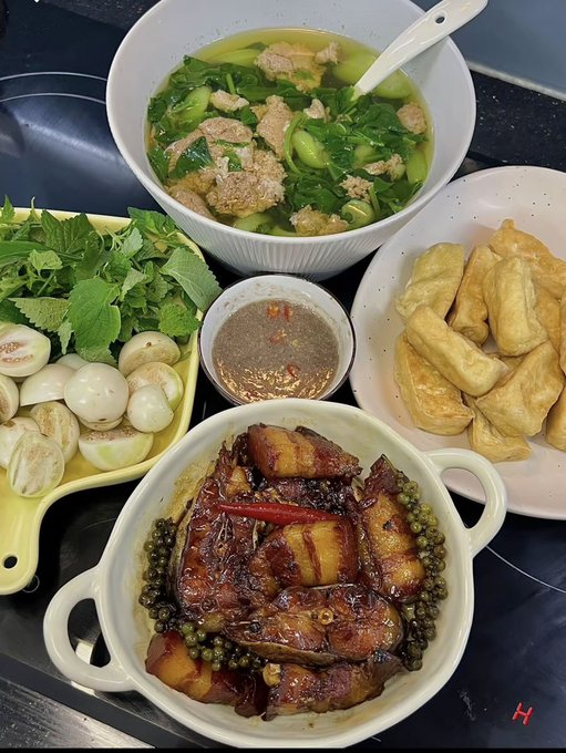 A spread of Vietnamese dishes including a bowl of soup with greens and pork, braised pork belly with peppercorns, fresh herbs with pickled garlic, fried tofu, and a dipping sauce.