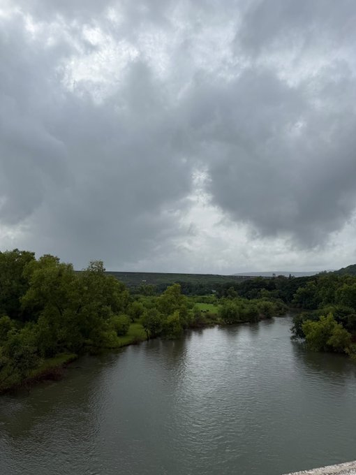 An overcast landscape featuring a wide river flowing through green, tree-covered hills under a cloudy gray sky.