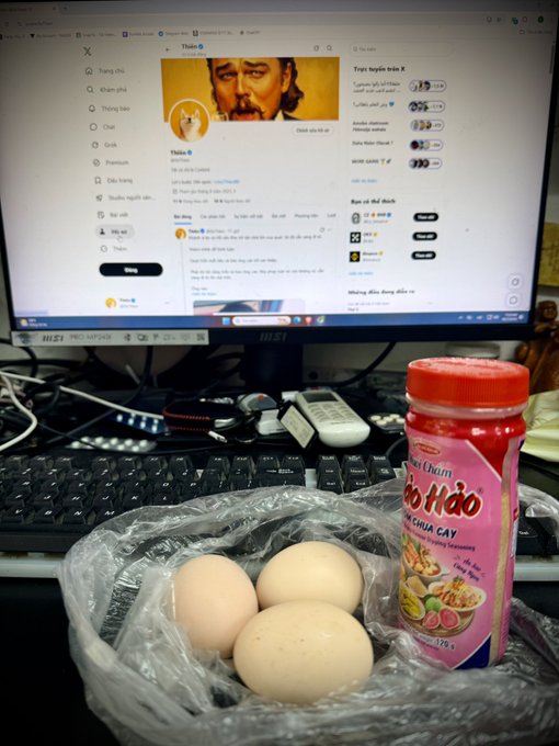 A desk scene showing a computer monitor displaying the X (Twitter) profile page of 'Thầu', with a keyboard, plastic bag of three eggs, and a bottle of Chao Hảo chili seasoning in the foreground.