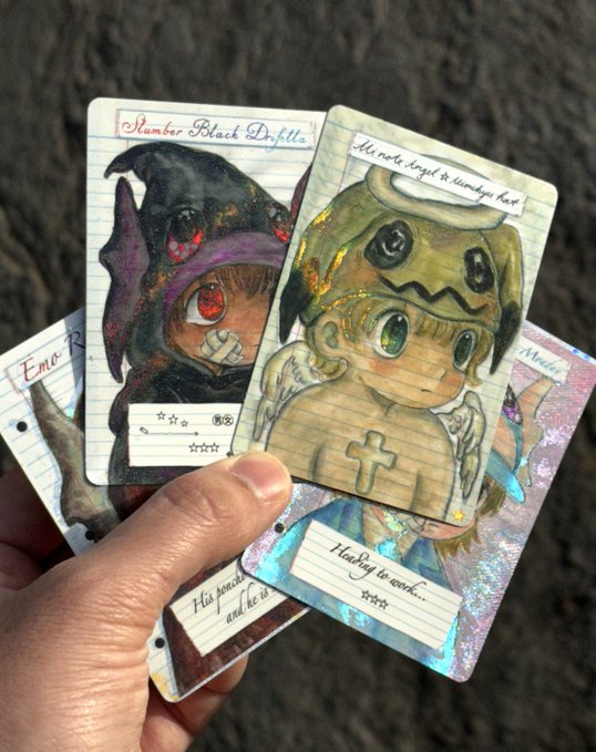 A hand holds three holographic illustrated character cards labeled "Slambar Black Dnfella," "It's not a angel it's starry eyed heart," and "His pain and He is... He's beg to work..." over a sandy beach. The artistic, character-themed note cards pair with the post's promotion of "mi note card packs" available at mons.shop.