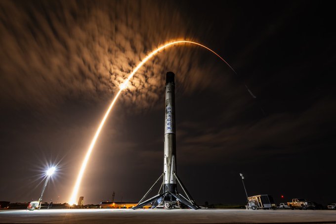 A SpaceX rocket stands on a launch pad at night, with a bright, curved trail of light arcing through the cloudy sky above it from a previous launch.