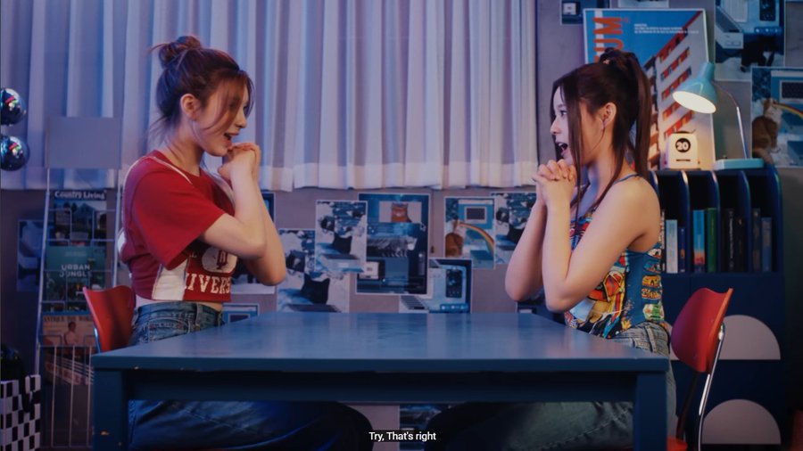 Two young women sit across from each other at a blue table in a retro-style room, clasping their hands together in excitement, with the subtitle 'Try. That's right.'