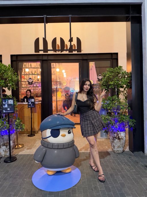 A woman posing with a peace sign next to a penguin mascot statue outside what appears to be a coffee shop, with the partial "mosa" sign above the entrance.