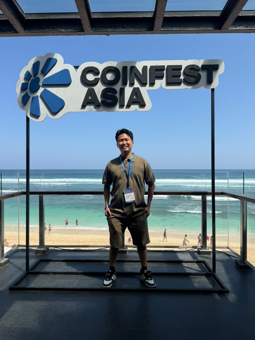A man wearing a lanyard with an event badge poses beneath a large 'COINFEST ASIA' sign overlooking a beach with turquoise water and surfers in the background.
