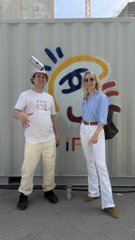 In this image, there are two people standing next to each other against a gray wall. One person is wearing a white shirt and hat, while the other person has blue pants and sunglasses. They appear to be posing for a photo together. The background of the image features a large mural on the wall that says "if you're still in Lisbon, make sure to stop by the GLiFS pop up at @opensea shipping container galleries before you leave help spray paint the mural, get a pair of GLiFSocks! Made in collab with my buddies at @apthcry". The text on the image suggests that they are in Lisbon and there is an art gallery nearby.