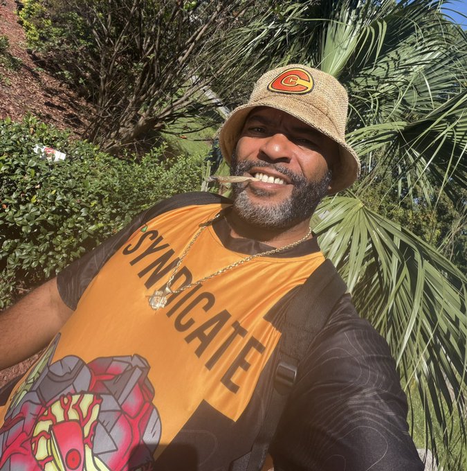 A selfie of a smiling man wearing a tan bucket hat with a 'C' patch and an orange 'SYNDICATE' t-shirt, with a lit cigar or joint in his mouth, set against palm fronds and shrubbery.