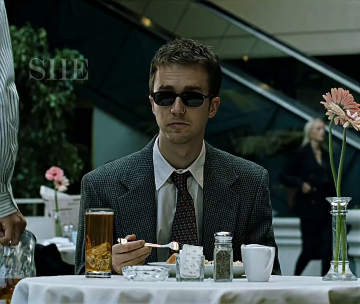 The image features a man sitting at a dining table, wearing sunglasses and holding a fork while eating food. The setting appears to be an indoor restaurant or cafe with various items placed around him, including a cup of beer, a wine glass, and some flowers on the table. There are also other people present in the scene, suggesting that this is a social gathering or event.