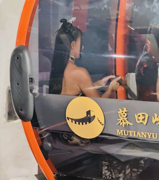 A photo taken through the window of a Mutianyu cable car, showing a woman inside, with the 'MUTIANYU' logo visible.