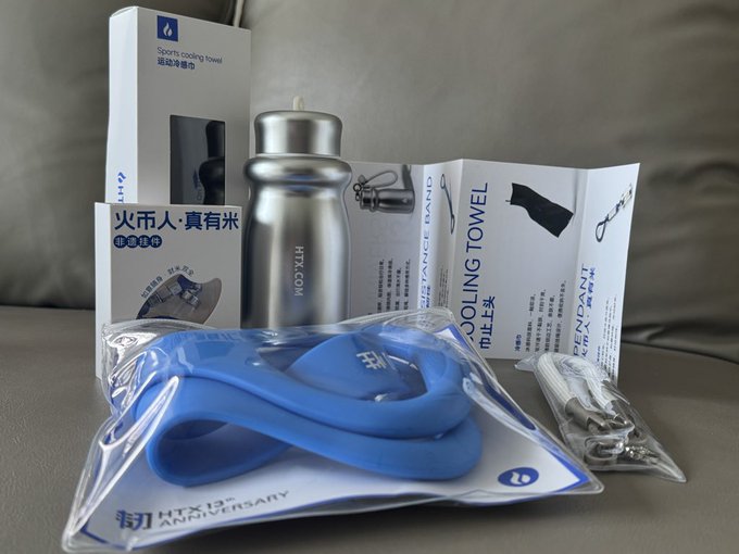 A collection of HTX-branded merchandise laid out on a couch, including a sports cooling towel, a metal water bottle, a resistance band, a pendant, a wristband, and a blue item in plastic packaging marked 'HTX 13th Anniversary'.