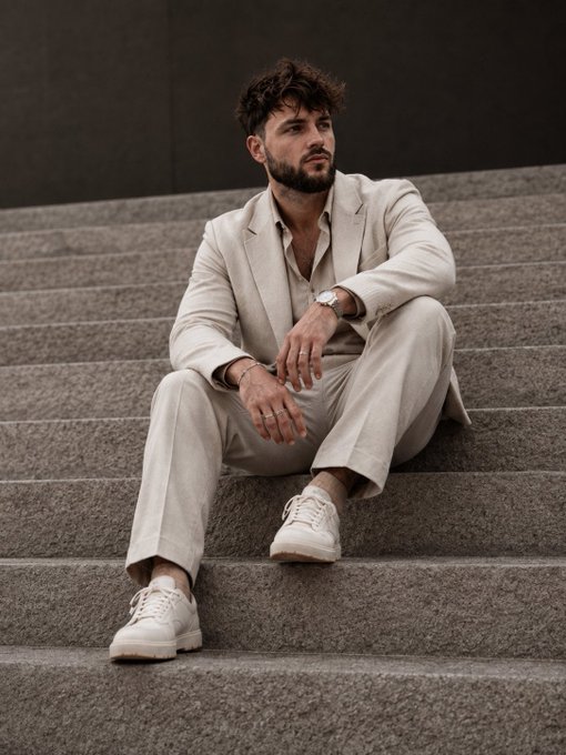 A man with dark curly hair and a beard, wearing a light beige suit and white sneakers, sitting thoughtfully on stone steps.
