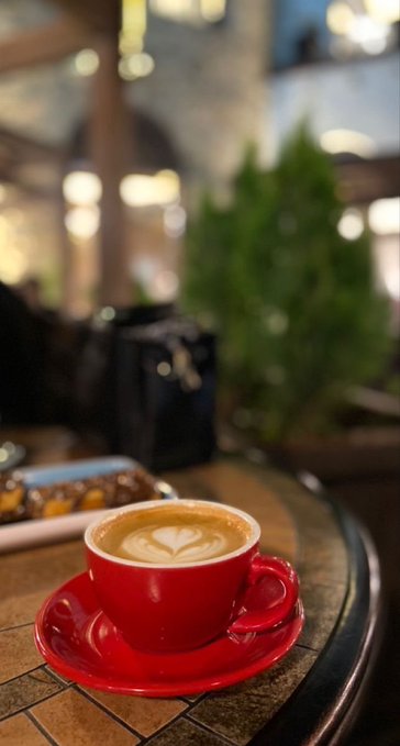 A red ceramic cup of cappuccino with heart latte art sits on a saucer on a cafe table.