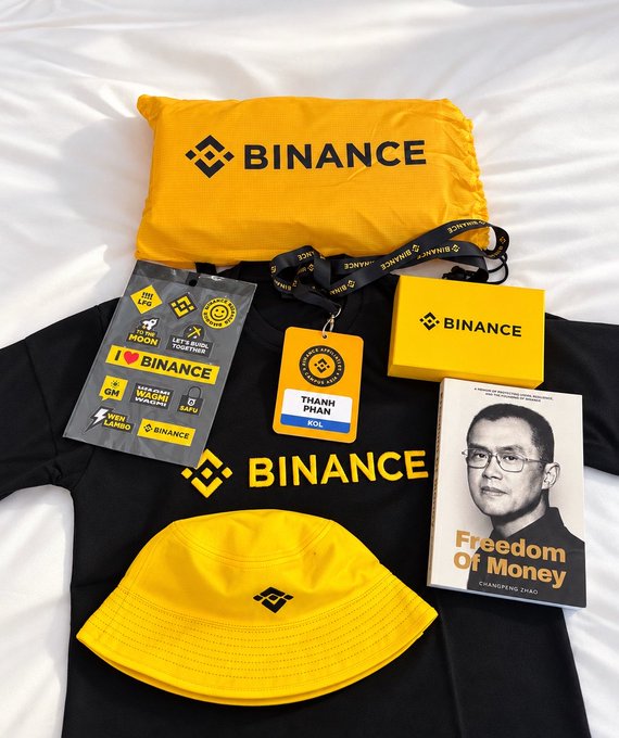 A flat lay of Binance merchandise including a yellow hat, lanyard with "THANH PHAN" name tag, stickers, t-shirt, and a copy of the book "Freedom of Money" by Changpeng Zhao (CZ).