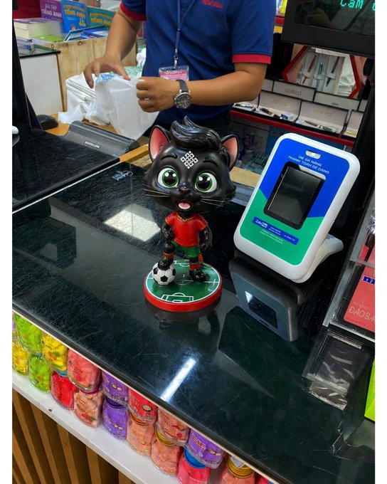 The image features a figurine of a cat, which appears to be a toy or decoration, placed on top of a black counter inside a store. The figurine has a soccer ball attached to its back, giving it an interesting and playful appearance. The scene also includes various items such as books and a computer mouse, suggesting that the store offers a diverse range of products for customers.

The image is accompanied by a caption that reads "Ritual: When AI, Web3, And Real-World Applications Come To Together". This phrase emphasizes the idea that the figurine represents the fusion of technology and real-world applications in today's society. The combination of these elements creates an intriguing visual narrative that invites viewers to consider how these different aspects might intersect and influence one another.