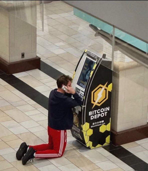 A young man kneeling in a mall in front of a Bitcoin Depot ATM, talking on a phone and appearing to embrace the machine.
