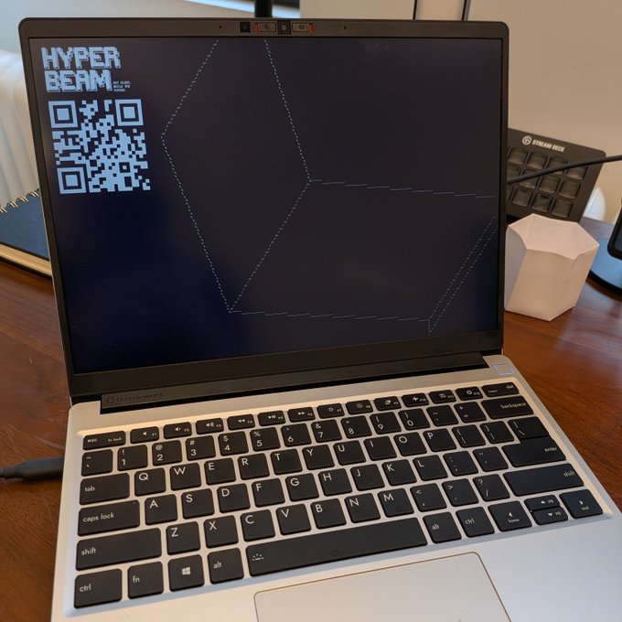 The image shows a laptop computer with its screen open, displaying a geometric shape or pattern that appears to be a code or a program. The laptop is placed on top of a wooden desk, which also has a keyboard and mouse nearby. The background features a windowed desktop computer setup, suggesting the presence of another computer in the room. The image includes text that reads "Running permawebOS", possibly indicating an operating system like Windows or Linux being used by the laptop's user.