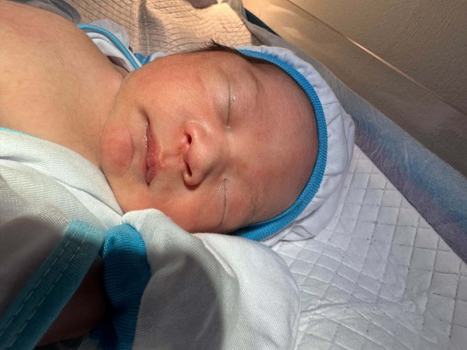 A newborn baby wearing a blue and white striped hospital cap sleeps peacefully on a quilted blanket.