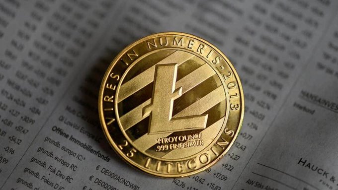 The image features a gold coin, possibly a Litecoin, placed on a newspaper or financial report page. The coin has a unique design that includes a vertical line running through its center. The text "It's the people's money" suggests that the coin is related to cryptocurrency and represents a digital currency with real-world utility. The coin is described as being cheap, reliable, and battle-tested for over a decade, indicating its long history in the market.