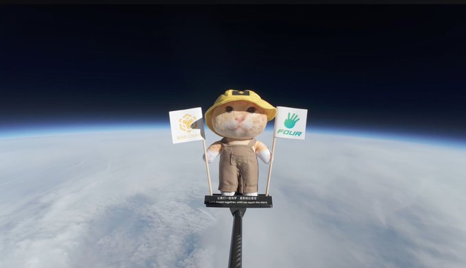 Image of a stuffed cat toy wearing a yellow hat and overalls, holding two small flags (one appearing to be Binance-related and one labeled 'FILBR'), suspended against a backdrop of Earth from high altitude.