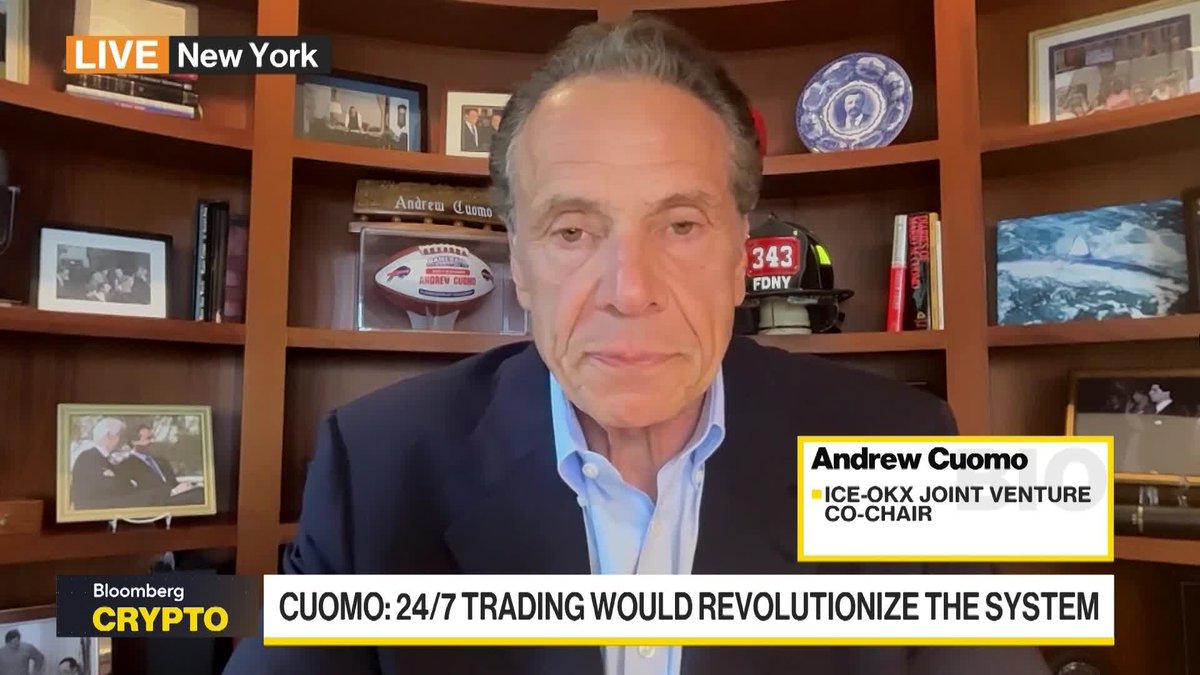 The image shows Andrew Cuomo, identified by the on-screen graphic as the ICE-OKX Joint Venture Co-Chair, appearing live on Bloomberg Crypto from New York. Behind him are shelves displaying personal memorabilia including a football labeled "Andrew Cuomo," an FDNY helmet marked "343," framed photos, and a decorative plate. The chyron reads "CUOMO: 24/7 TRADING WOULD REVOLUTIONIZE THE SYSTEM," aligning with his post advocating for clear blockchain regulations so the U.S. can lead in market innovation rather than fall behind.