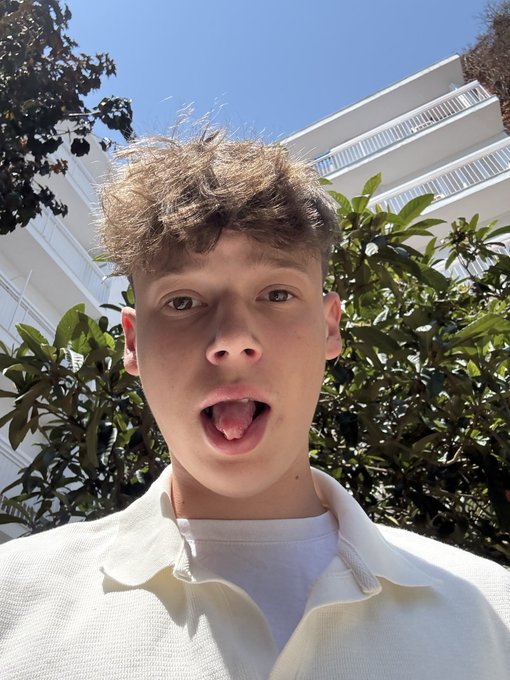 Close-up selfie of a young man with curly brown hair wearing a cream-colored shirt, standing outdoors with green foliage and a white building in the background.