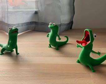Three little green toy dinosaurs posed on a wooden surface, with one in the foreground with its mouth wide open as if roaring, while two others stand in the background near a striped curtain.