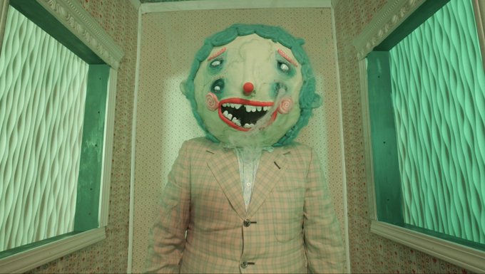A creepy figure wearing a plaid suit jacket stands in a greenish room with wavy-patterned windows. Its oversized head is a grotesque clown-like mask with a wide grinning mouth showing sharp teeth, a red nose, and multiple eyes.
