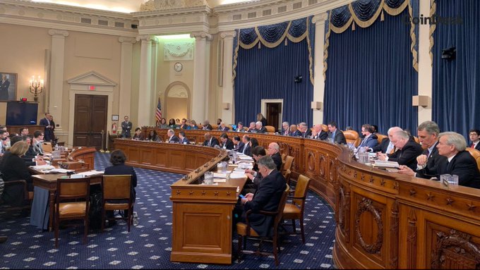 The image shows a congressional hearing or committee meeting taking place in an ornate government chamber, with numerous lawmakers and staff seated around a curved wooden dais and at tables, with an American flag visible in the background. This setting reflects the formal legislative process behind the House Ways and Means Committee's upcoming vote on the Digital Asset Tax Certainty Act, illustrating the institutional setting where such crypto tax legislation is being deliberated.