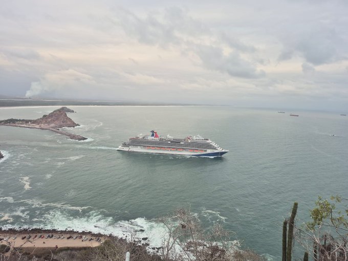 The image features a large cruise ship sailing across a body of water, possibly an ocean or sea. The ship appears to be moving towards the right side of the frame. In the background, there are several other boats visible at various distances from the main ship. The sky above is cloudy and gray, suggesting that it might be a slightly overcast day.