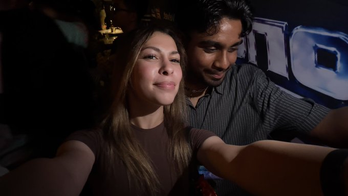 A selfie-style photo of a young woman in a brown shirt and a young man in a dark striped shirt posing together in a dimly lit setting with a blue banner in the background.