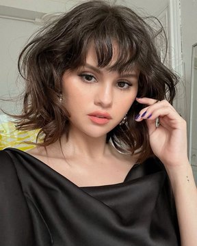 A portrait of a young woman with shoulder-length wavy dark hair and bangs, wearing a black satin top, posing with her hand near her face.