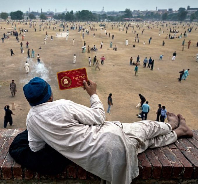 A man in traditional South Asian attire with a blue turban lies on a brick ledge, smoking and holding up a 'TMP PASS' card (No. 0717, The Mughal City) while looking out over a vast field filled with many cricket players, likely in Lahore, Pakistan.