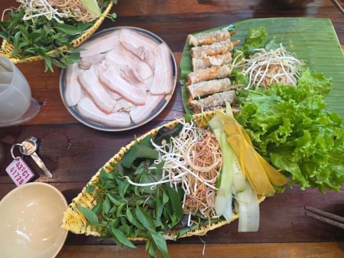 A wooden table set with Vietnamese food, featuring fresh herbs and lettuce on banana leaves, sliced pork, and rolled spring rolls, with a hotel key tag labeled 'R38CT 11728' visible.
