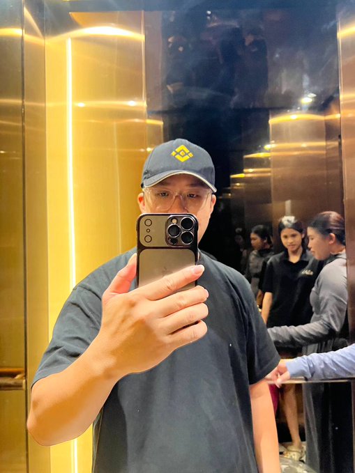 A man wearing a navy cap with a yellow logo takes a mirror selfie with an iPhone in a gold-toned elevator lobby, with other people visible in the background.