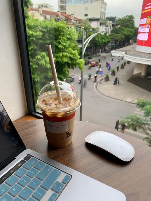 An iced coffee sits on a wooden desk beside a MacBook with a blue keyboard cover and a white Apple Magic Mouse, overlooking a bustling city street below from a high-rise window. The post's mention of skipping a Da Lat trip due to laziness mirrors this cozy indoor scene, where procrastination has traded adventure for a relaxed caffeine fix with a street view.