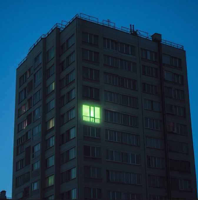 Nighttime photograph of a tall apartment building with one apartment window illuminated in bright green, glowing like a pixel art character.