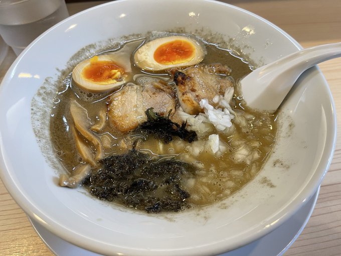 A close-up photo of a bowl of Japanese ramen featuring soft-boiled egg halves with orange yolks, chashu pork, mushrooms, seaweed, and rice in a dark broth, served with a white ceramic spoon.